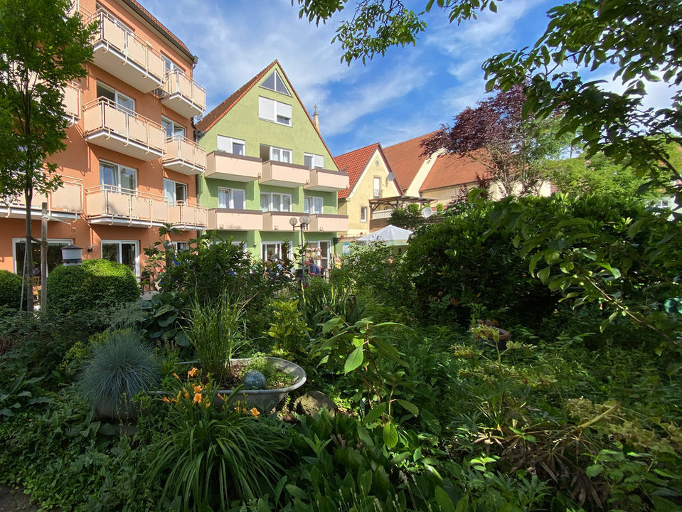Blick durch das Grün im Garten auf die zwei Gebäude des Gasthofs und Hotels. das rechte Gebäude ist grün und hat ein Spitzdach und das linke Gebäude ist rot/orange und hat ein flaches Dach. Beide Gebäude haben mehrere Stockwerke mit Fenstern und Balkonen.