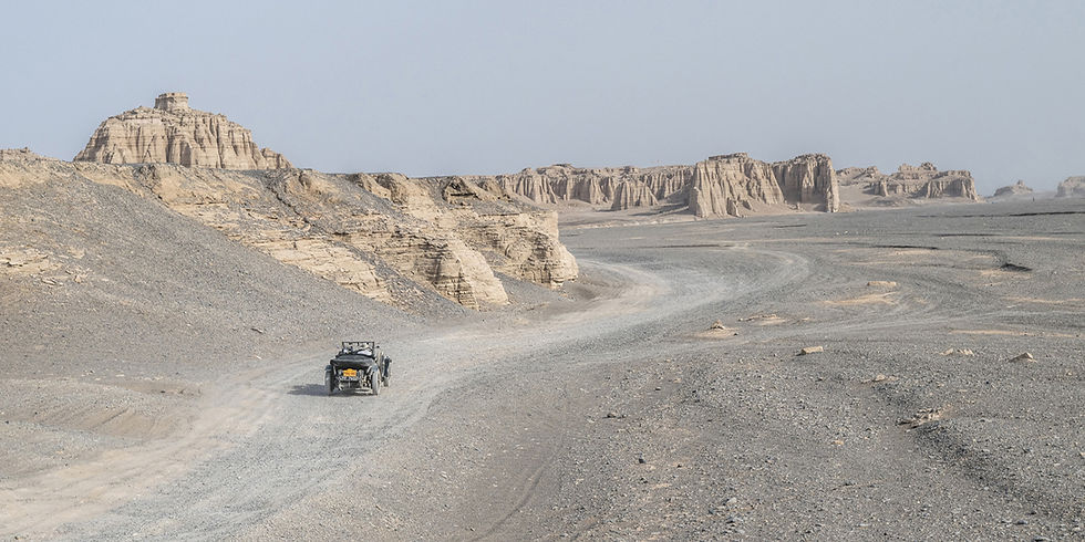 Vintage Bentley car driving along a remote gravel track through a vast, barren desert landscape surrounded by dramatic rock formations.
