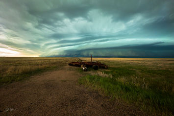 A blueish-green storm containing a large shelf cloud is in the distance, hovering over a field containing some old farming equipment in the foreground.