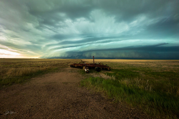 A blueish-green storm containing a large shelf cloud is in the distance, hovering over a field containing some old farming equipment in the foreground.