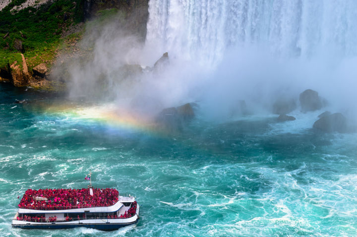 Niagara Falls is visible on the right side of the frame, with mist causing a rainbow taking up the frame. A tour ship is seen going into the mist. Land is visible on the left side of the frame.