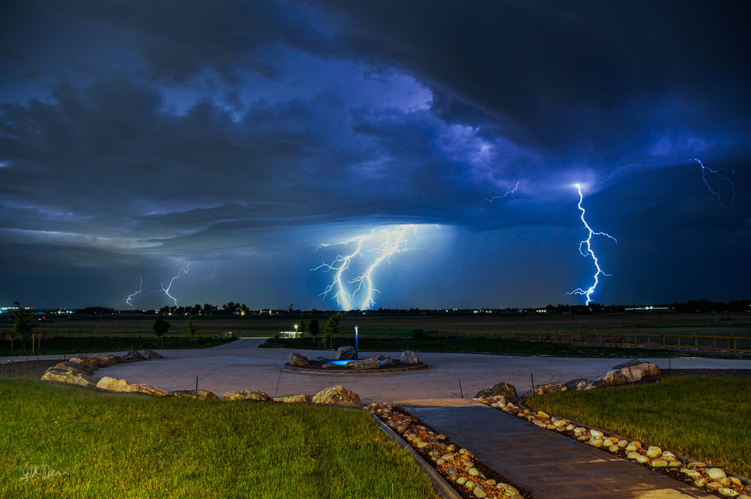 A storm with multiple blue-ish lightning bolts is in the distance. A green field and stone paths are in the foreground.
