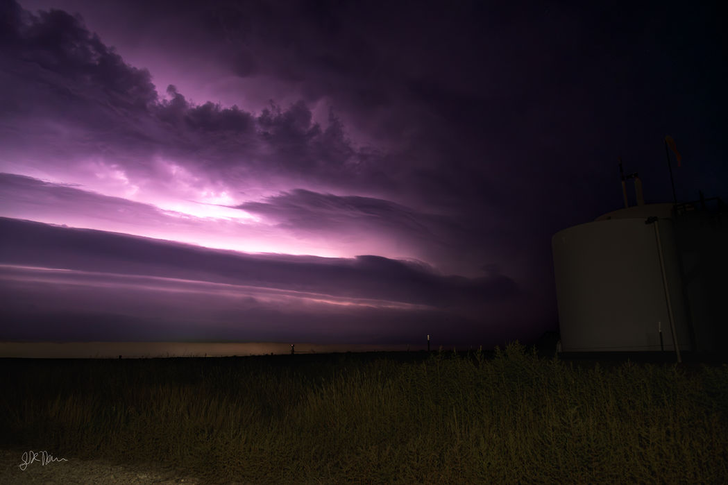 A night sky is dramatically illuminated from within by intra-cloud lightning, casting an intense violet and magenta glow behind stacked, horizontal storm clouds. In the foreground, tall wild grasses and a large silhouette of a storage tank or silo frame the right edge against the glowing horizon.