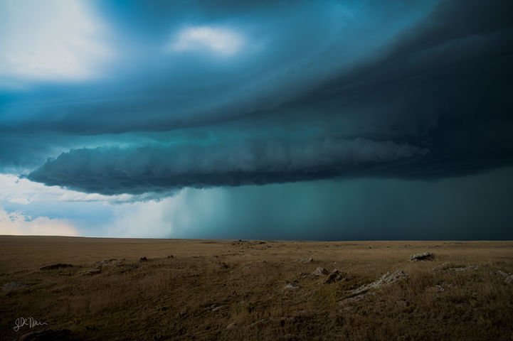 A massive, dark supercell storm towers over a vast, rocky plain under a heavy, layered shelf cloud. Beneath the shelf, an intense, brilliant teal and cyan precipitation core drops a heavy wall of rain and hail to the distant horizon, contrasting sharply with the barren, light-brown grassy landscape in the foreground.
