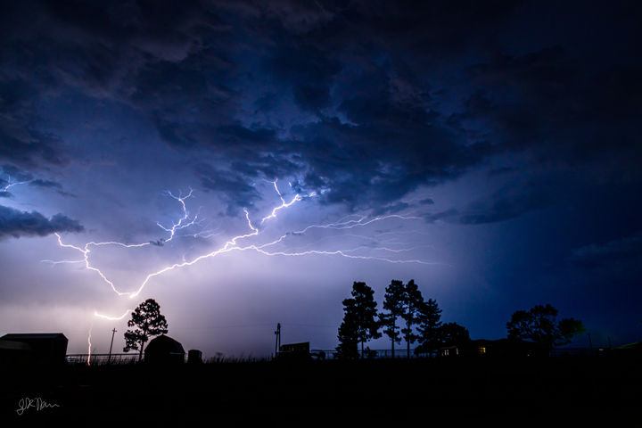 A lightning strike reaching out from the ground over the sky and a farm with some trees on the bottom.