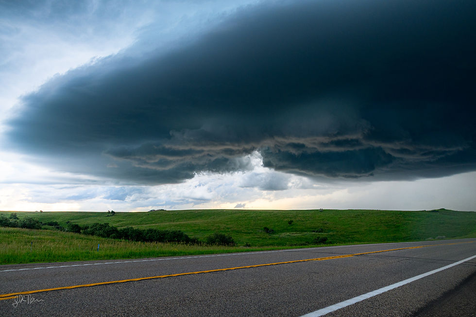 A dramatic storm cell with a large, rotating wall cloud and a visible RFD clear slot looming over a rural highway and rolling green hills.