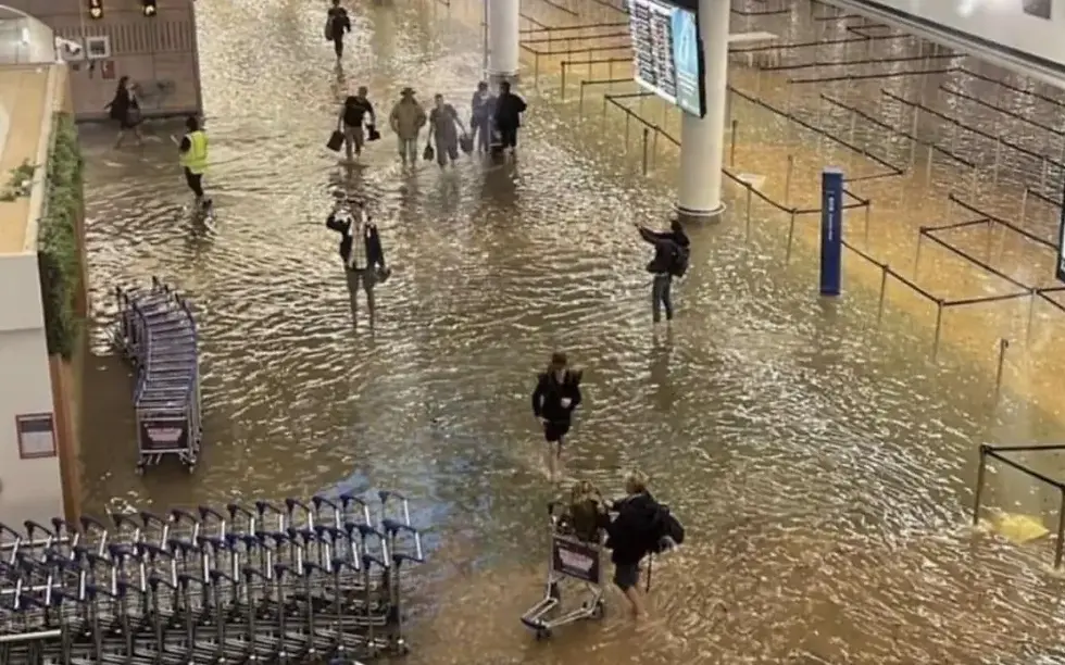 People standing inside Auckland Airport terminal in brown ankle-depth flood water, with nearby trolleys