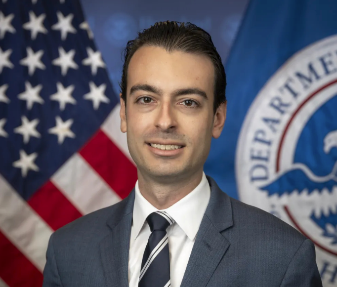 Smiling man in suit and tie stands in front of U.S. flag and Homeland Security flag. Formal setting, patriotic decor.