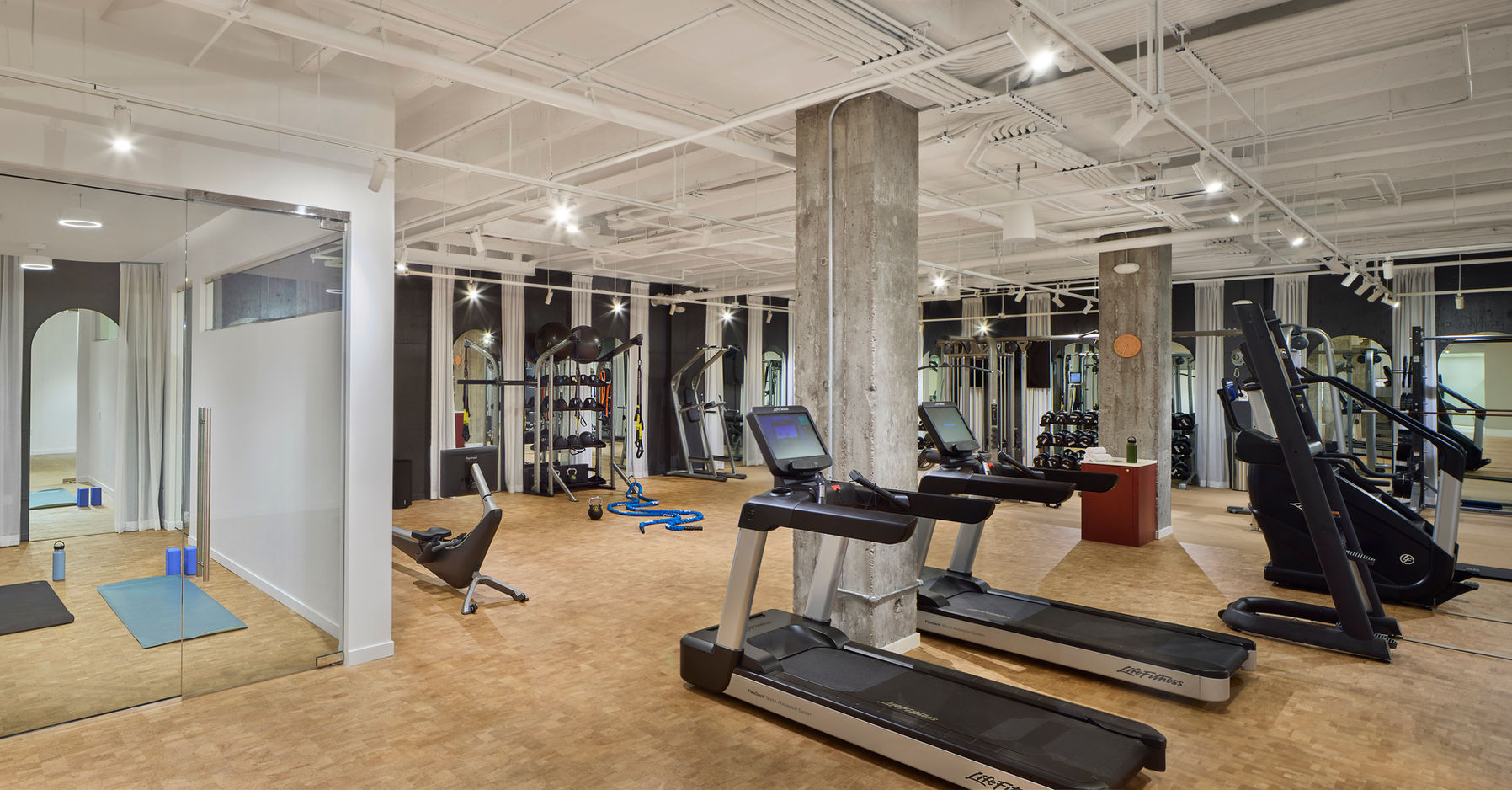 Modern commercial gym interior featuring a spacious, well-lit fitness area with treadmills, elliptical machines, and strength training equipment. Large mirrors line the walls, enhancing the open feel of the space. A concrete pillar stands centrally, adding an industrial design element. Rubberized flooring provides durability and safety for workouts. In the corner, a glass-enclosed yoga or stretching room offers a private wellness space.