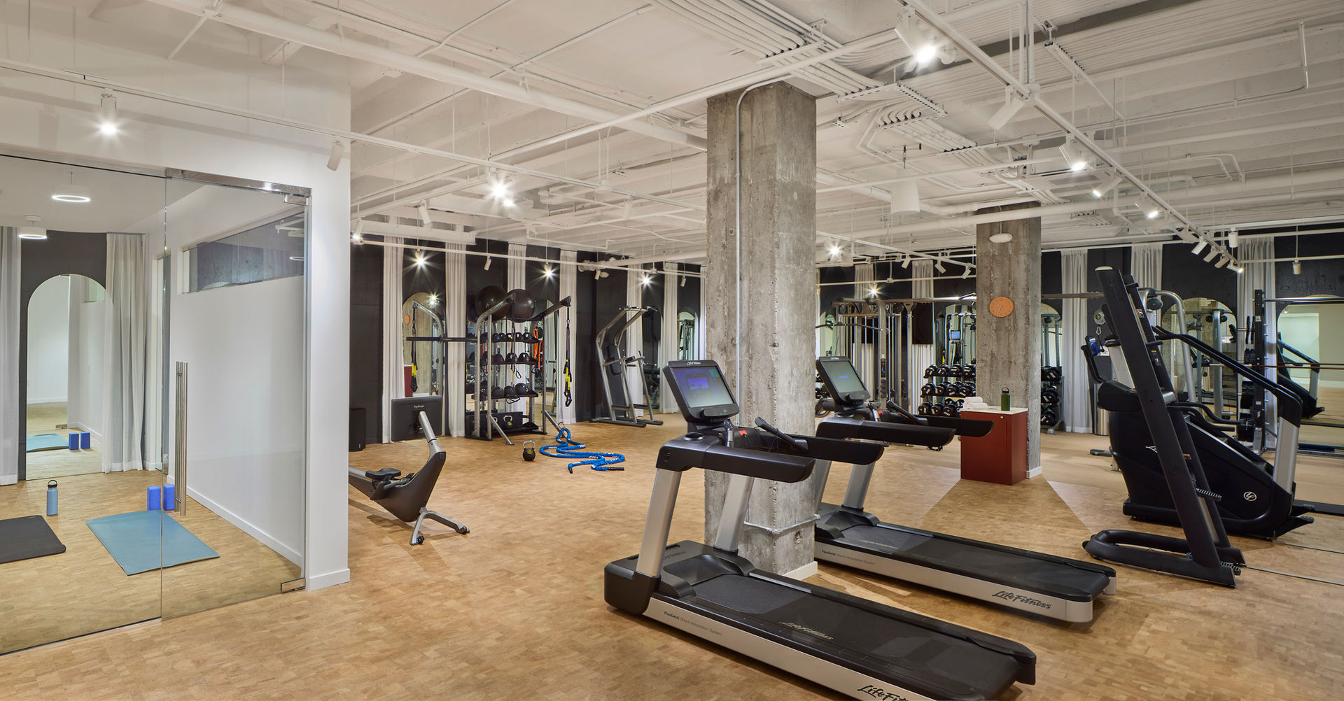Modern commercial gym interior featuring a spacious, well-lit fitness area with treadmills, elliptical machines, and strength training equipment. Large mirrors line the walls, enhancing the open feel of the space. A concrete pillar stands centrally, adding an industrial design element. Rubberized flooring provides durability and safety for workouts. In the corner, a glass-enclosed yoga or stretching room offers a private wellness space.