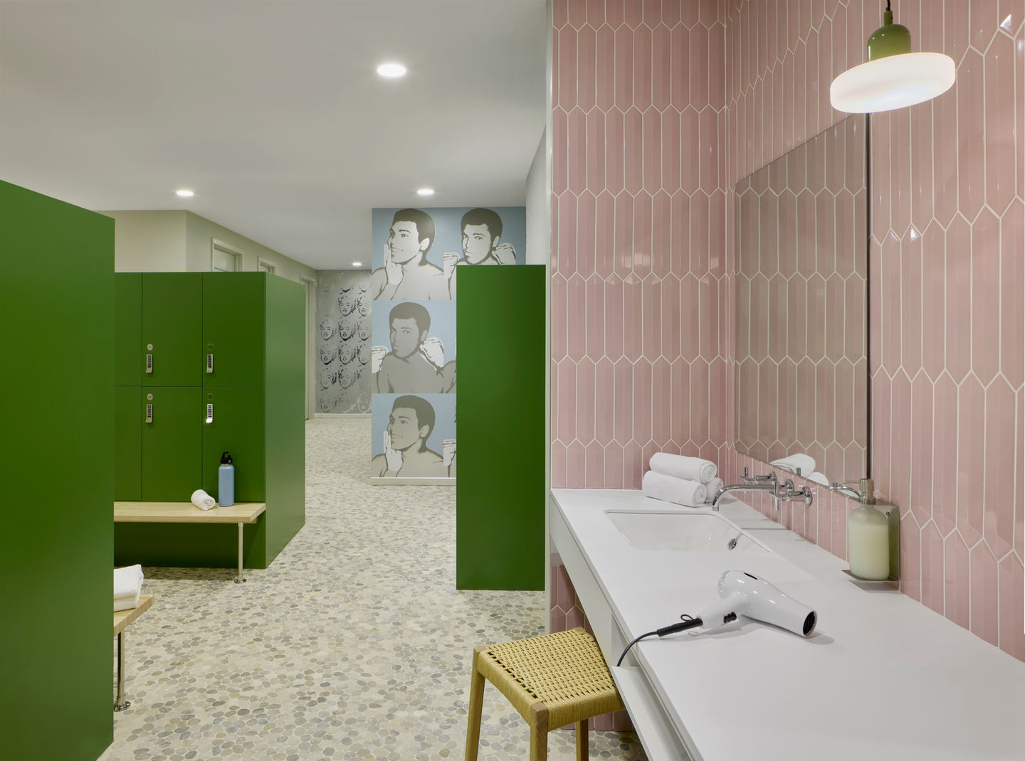 Contemporary gym locker room featuring green lockers, pink geometric wall tiles, and a long white vanity with a hairdryer, candle, and rolled towels. A wooden stool sits beneath the counter, and mosaic-style flooring adds texture throughout the space. The background reveals a wall mural of stylized male portraits, enhancing the modern aesthetic. The space is brightly lit and thoughtfully designed for comfort, grooming, and post-workout relaxation.
