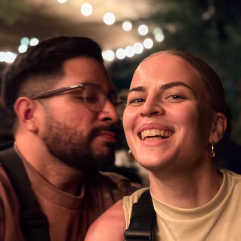 A woman smiling to camera and man giving her a cheek kiss.
