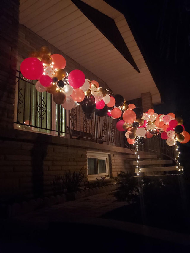 A pink and white balloon garland with string lights along a porch railing at night