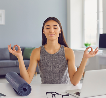 A Woman sitting at her desk with her eyes closed in a meditative pose. A computer to her left and exercise mat to her right.