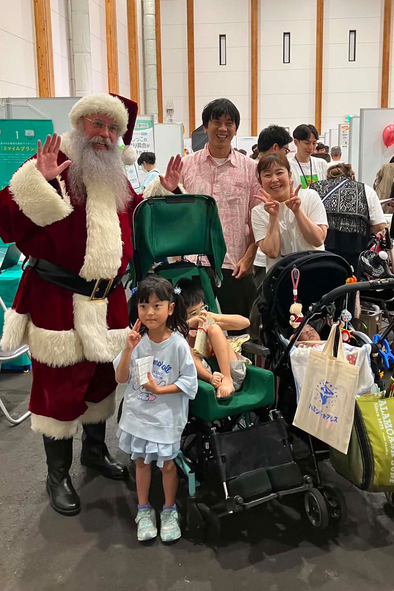 屋内イベント会場で、サンタ姿の男性と家族がベビーカーの前で笑顔で手を振り、子どもたちがピースしている。