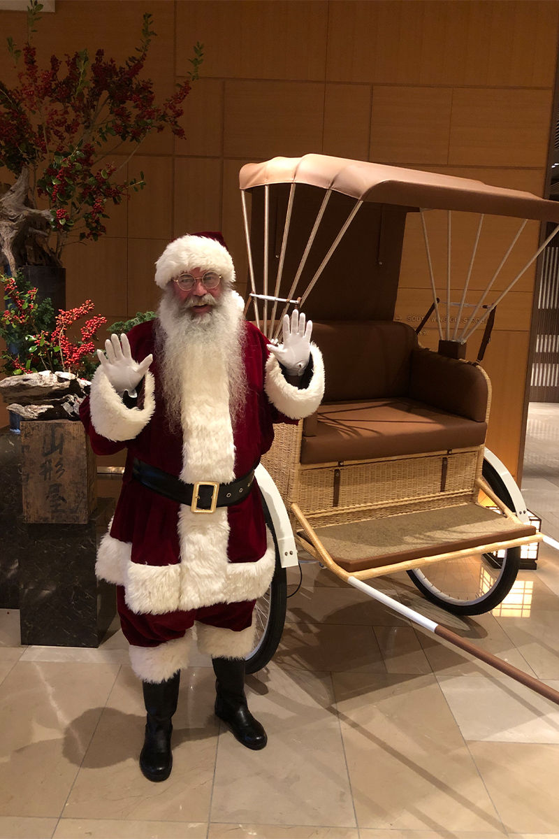 サンタクロース姿の男性がリヤカーの前で手を挙げている。背景にクリスマスの装飾があり、温かい室内の雰囲気。