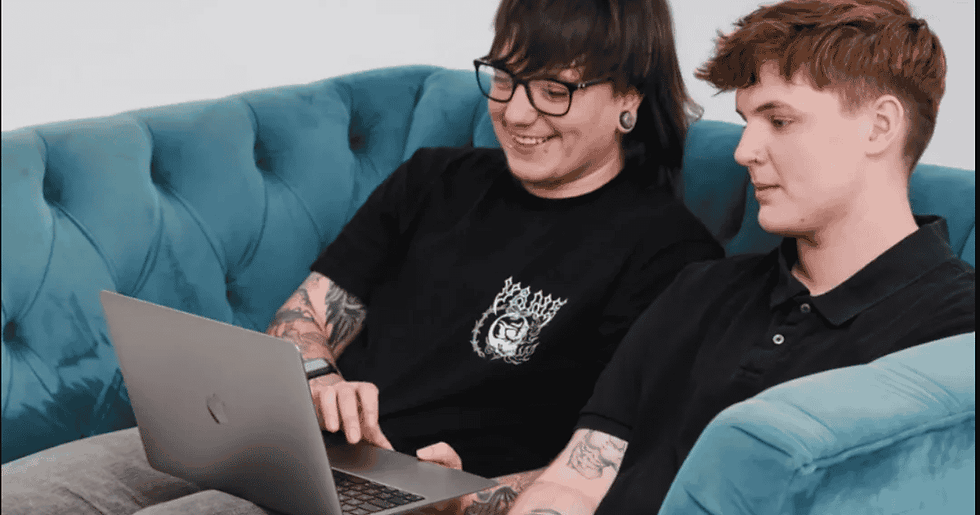 Two people sit on a blue couch, smiling at a laptop. Both have tattoos and wear black shirts. Relaxed, casual setting.