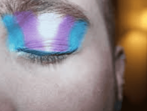 Closed eye with bold makeup in blue, purple, and pink hues. Blurred warm lighting in the background creates a cozy ambiance.