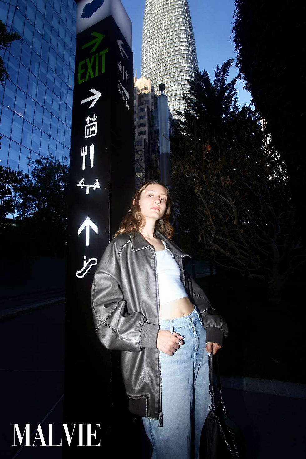 Model leaning on a tall exit sign in an oversized leather jacket, a skyscraper behind