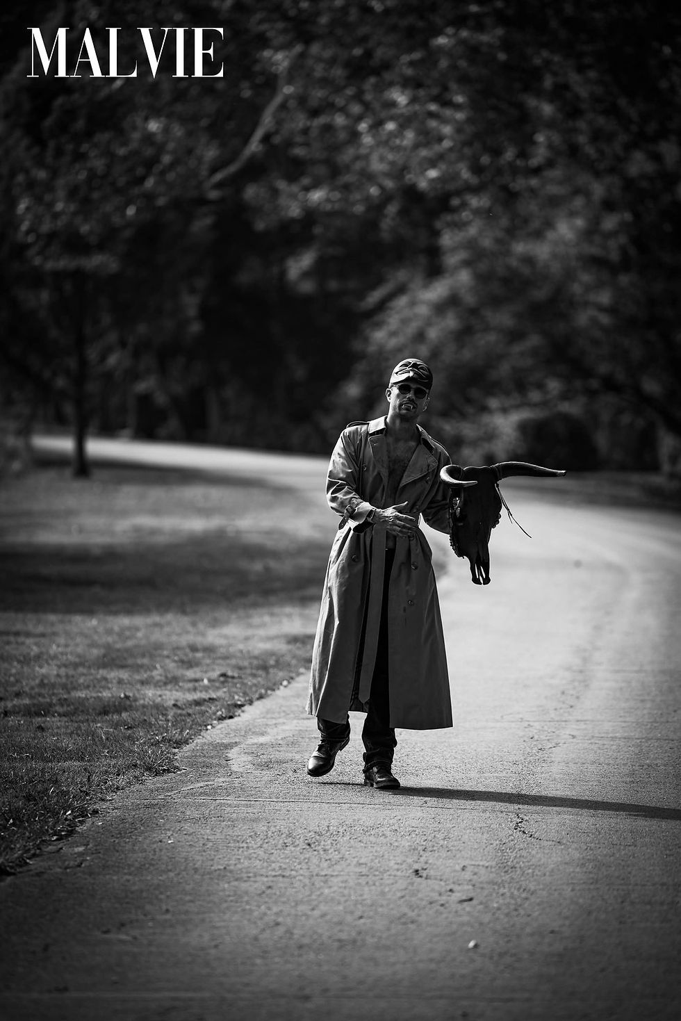 Figure in a trench coat on a tree-lined road with a black bird in hand
