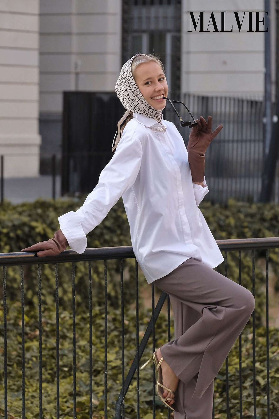 Model laughing as she leans on a railing above a hedge, in a headscarf, white shirt and taupe trousers
