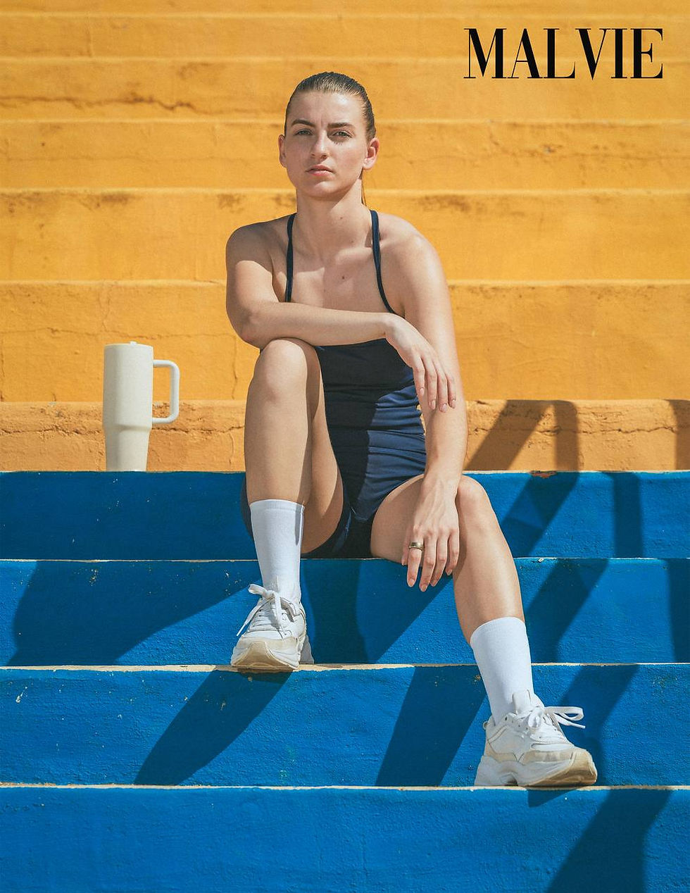 Model sitting on cobalt-blue steps in a navy romper, white socks and trainers, a white tumbler beside her