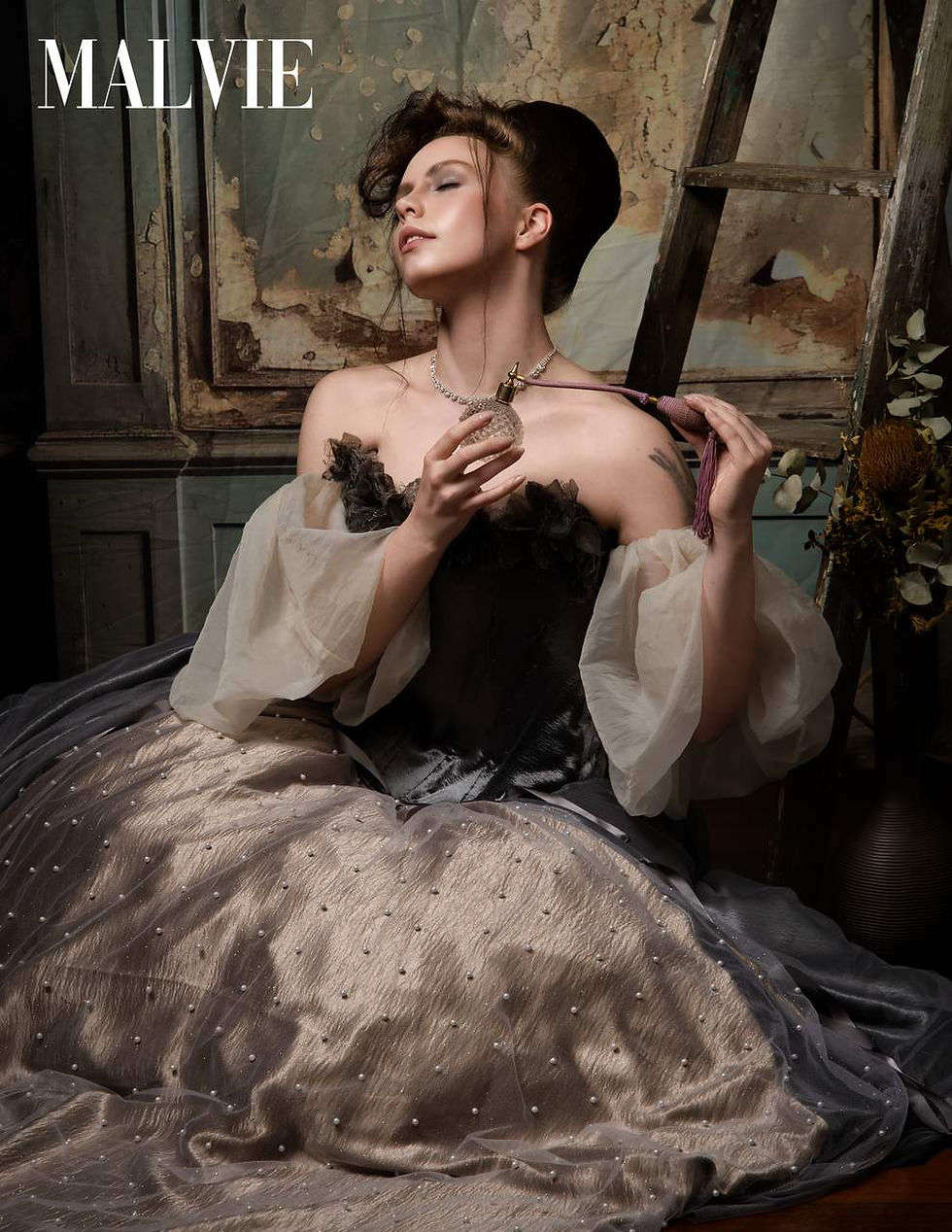 Model seated in a pewter satin gown with sheer puffed sleeves, eyes closed, against a peeling wall