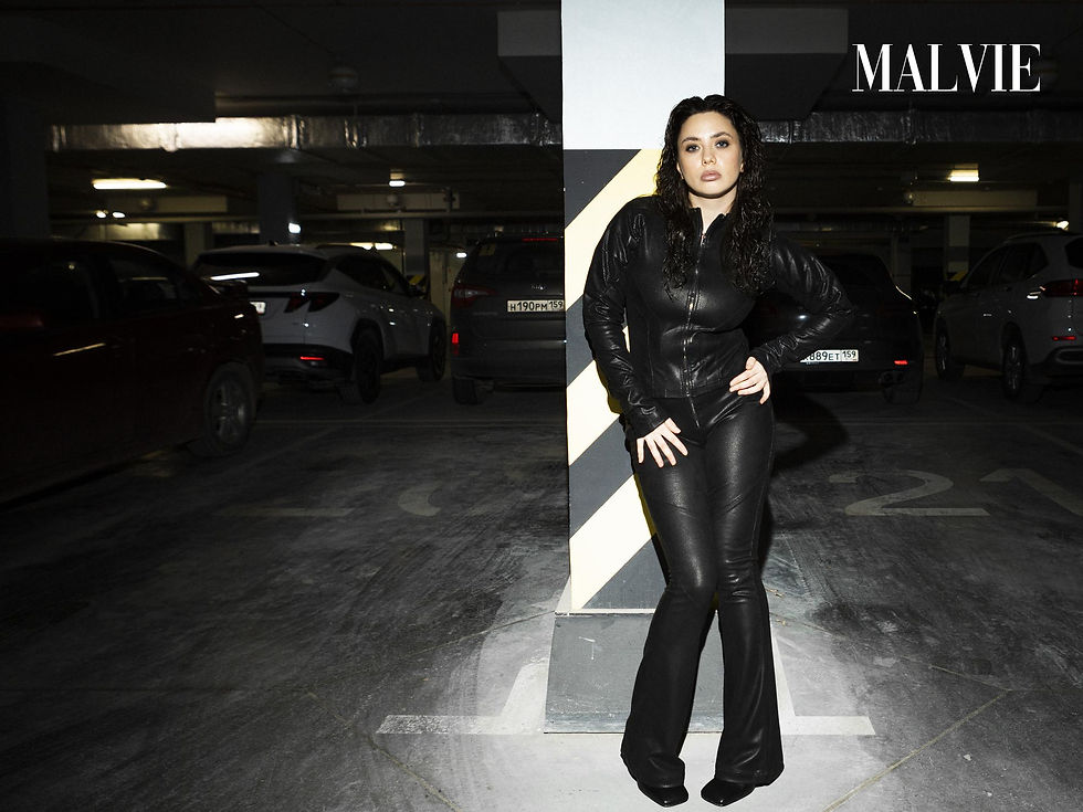 Model in black leather, hand on hip, leaning on a white pillar among parked cars