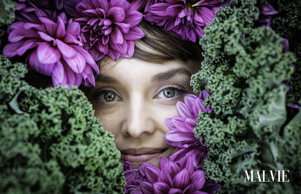 Model smiling softly, her face framed by curly green kale and a few purple flowers