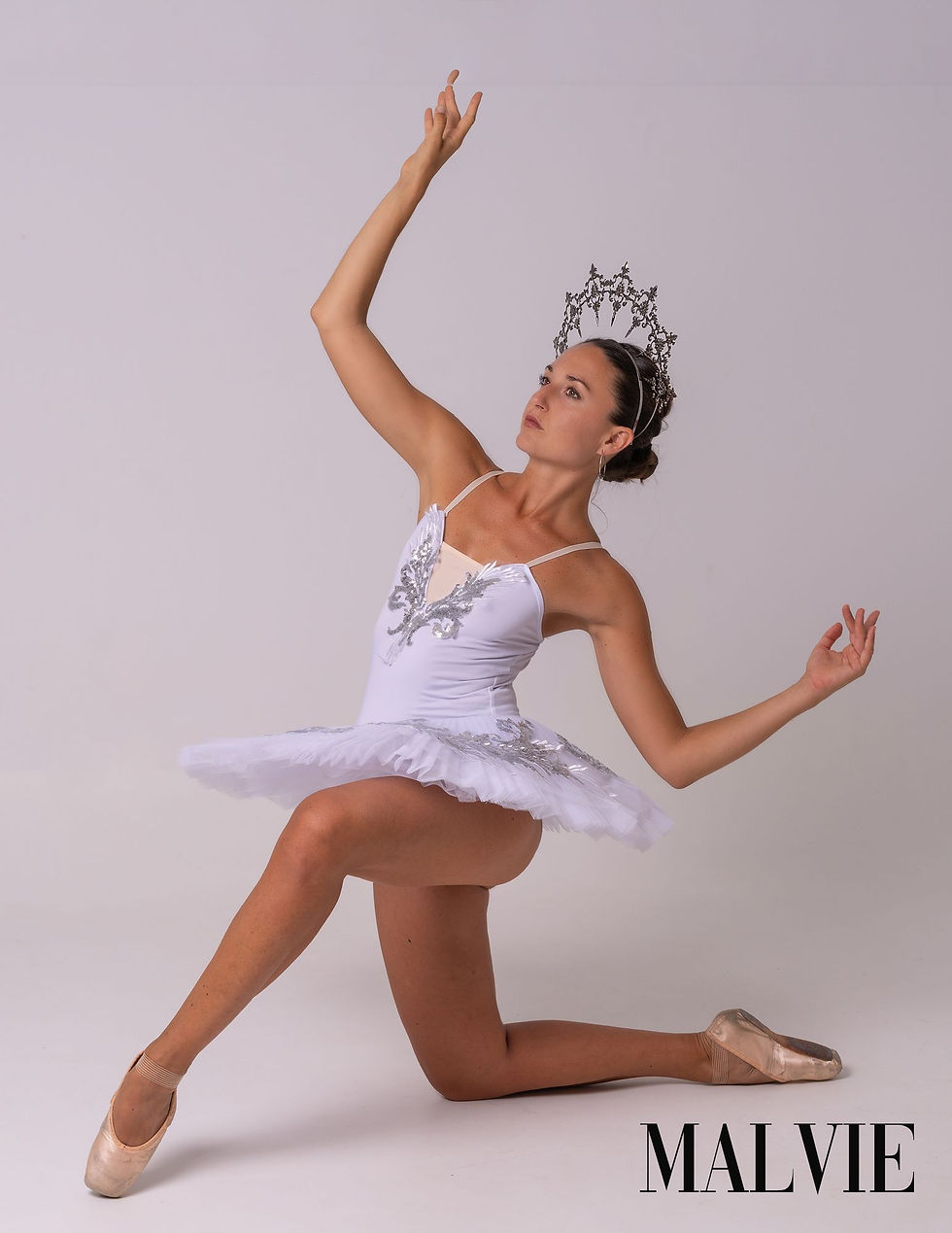 Model in a white tutu and silver tiara kneeling on one knee, one arm raised, in pointe shoes