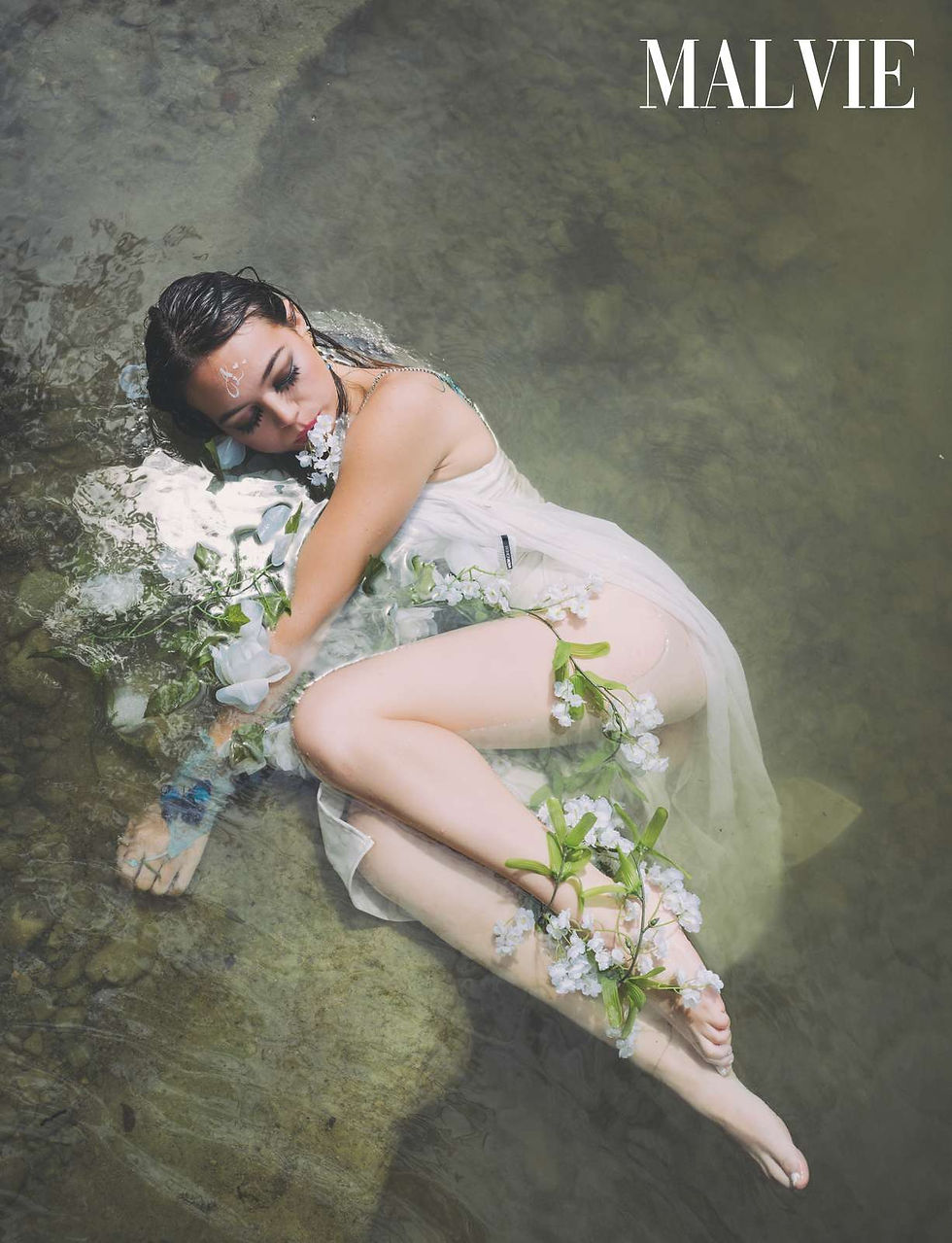 Model curled on her side on the riverbed among white blossoms