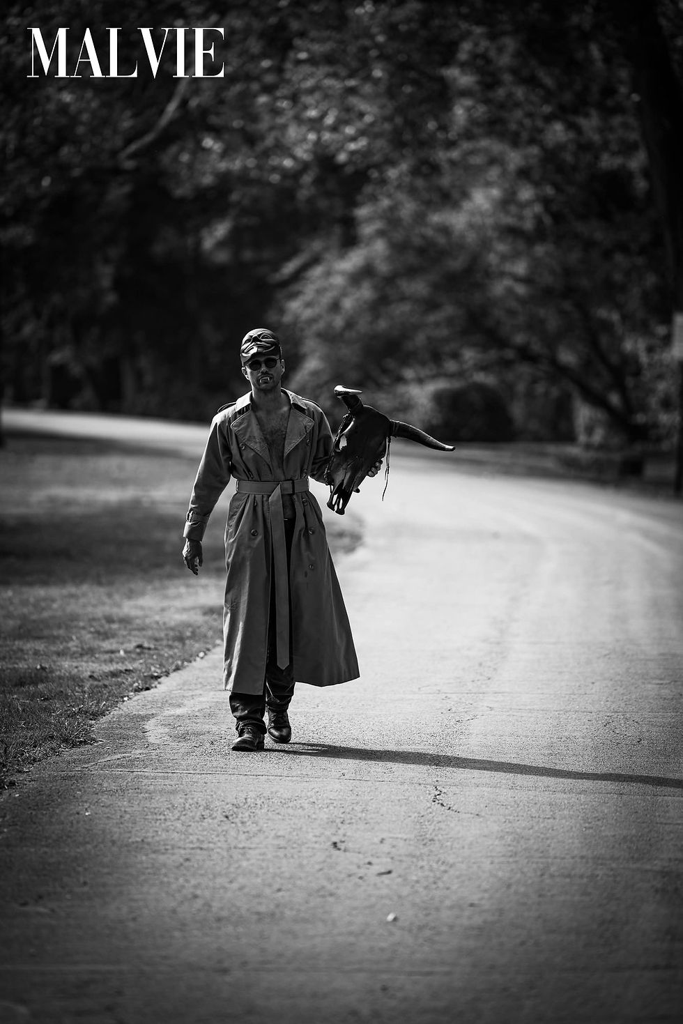 Figure in a long trench coat carrying a black bird on a tree-lined road