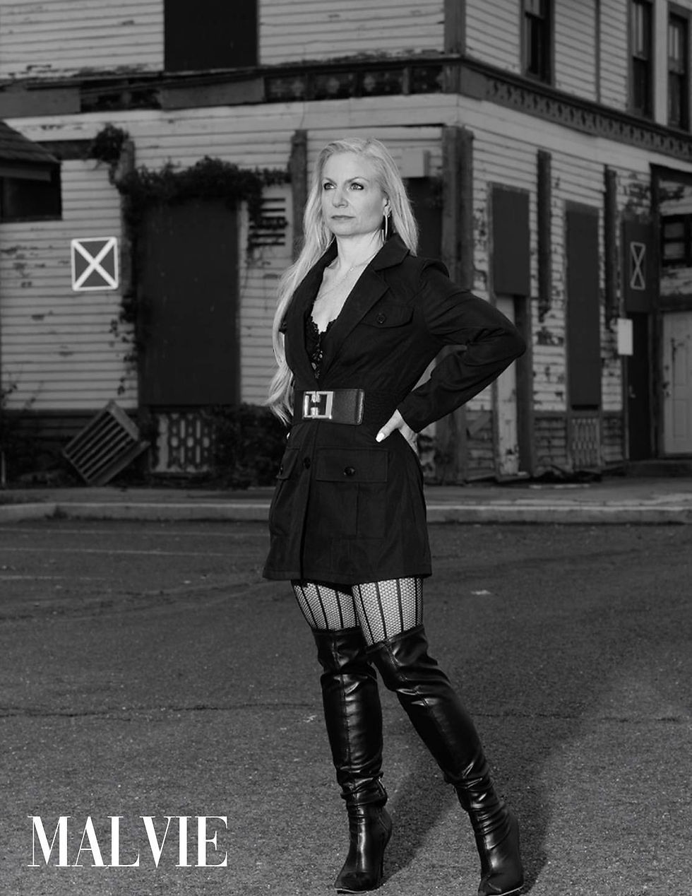 Model with a hand on her hip in a belted black coat dress in front of an old wooden building