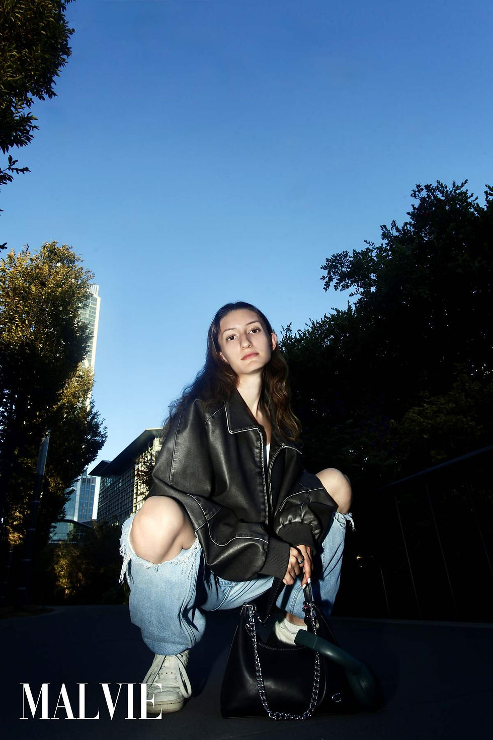 Model crouching on her heels in a leather jacket and ripped jeans under a blue sky