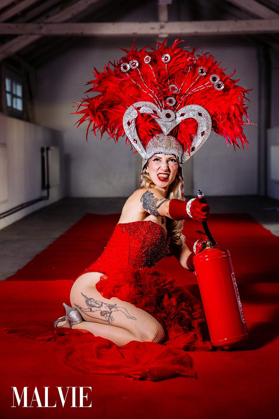 Model kneeling on the red carpet beside a red fire extinguisher, laughing, in a red sequinned bodysuit