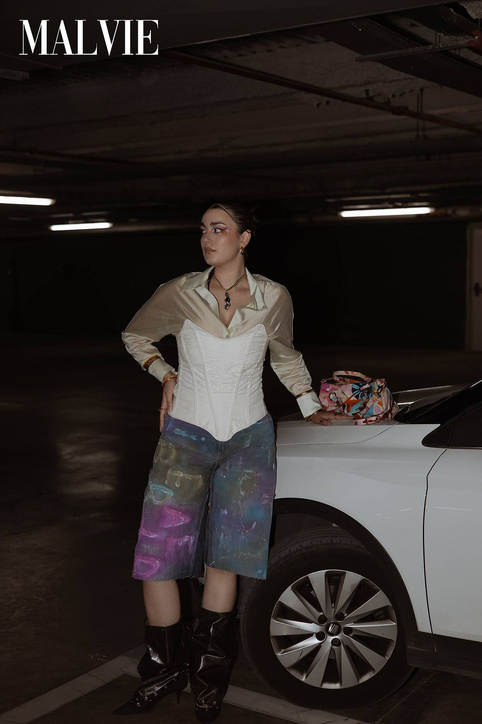Model standing with a hand on her hip beside a white car in painted Bermuda shorts and black boots