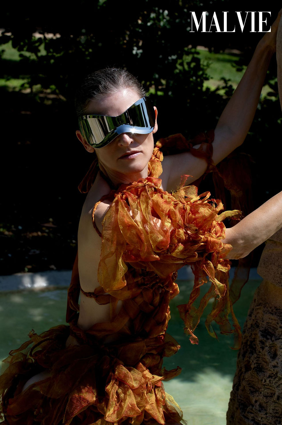 Guiomar Alfaro Pablos in a rust feathered dress and mirrored visor sunglasses by a garden fountain