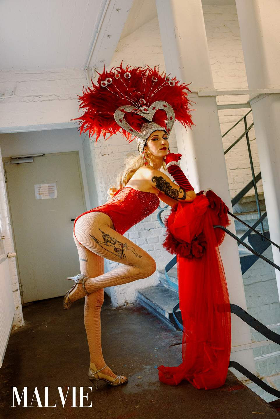 Model posing on one leg in a red bodysuit and long tulle train against a pillar by the stairs