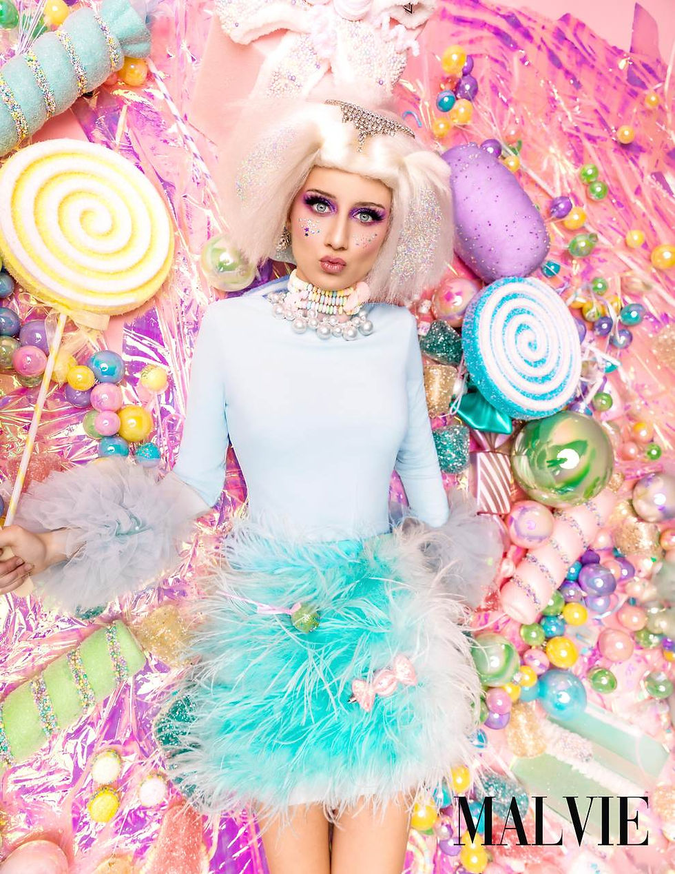 Model in a platinum wig and powder-blue top with a mint feather skirt, a giant lollipop beside her