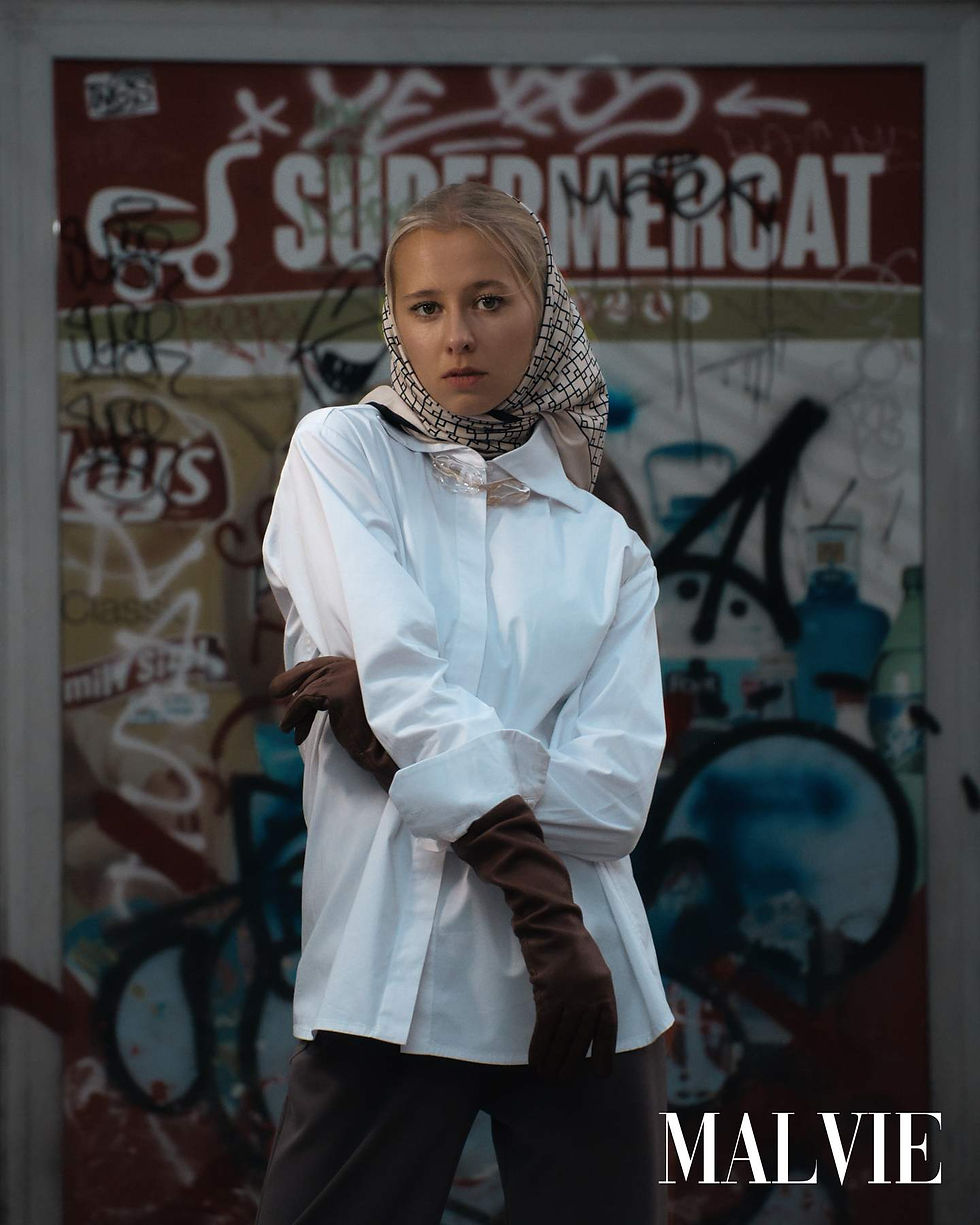 Vianne in an oversized white shirt, patterned headscarf and long brown gloves in front of a graffiti-covered shop sign