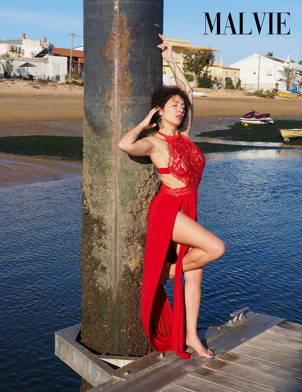 Model in red lace and a red slit skirt leaning against a rusted post on a jetty, houses behind