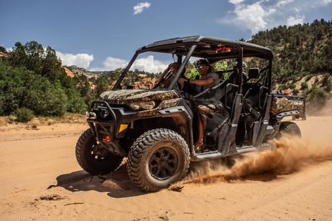 Two individuals riding fast in the front seat of a four-person camo UTV driving through sand.