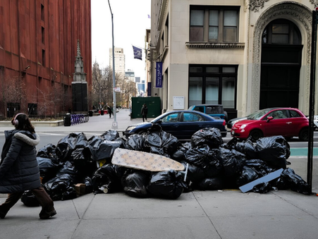 A woman wearing a heavy puffer coat and pink headphones walks by a large pile of trash bags on a city street