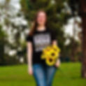 Jessica smiles at the camera while holding a bouquet of sunflowers at her hip. She's wearing a black shirt with white and colored text saying, "Disability rights are human rights," along with disability symbols.