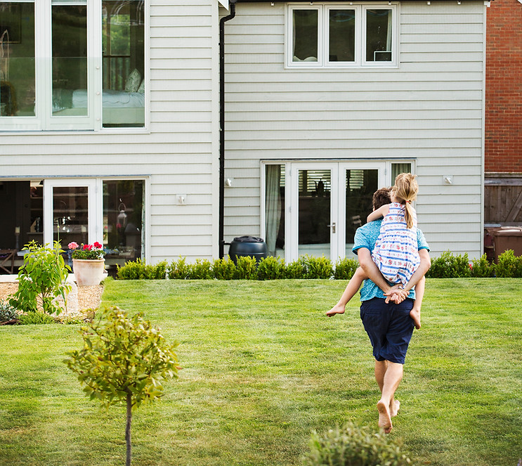 Father carrying daughter toward modern family home with large backyard, flat fee MLS real estate listing in Quebec