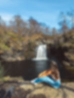 Woman in jeans sits by a river, gazing at a waterfall surrounded by trees under a clear blue sky, conveying serenity.