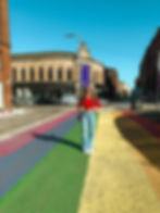Woman in red top stands on a rainbow-painted street. Historic buildings surround her under a clear blue sky. She smiles, wearing sunglasses.