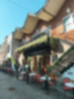 Outdoor cafe with red chairs and tables on a cobblestone street. Greenery drapes over a brick building labeled "The Gardener." Blue sky above.