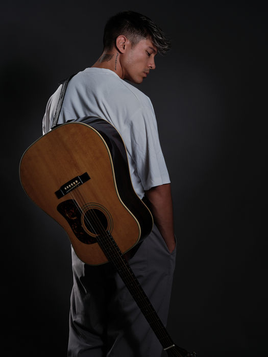Mann von hinten mit Gitarre um die Schultern. Das Gesicht wird im Profil gezeigt. Weisses T-Shirt und graue Hose.