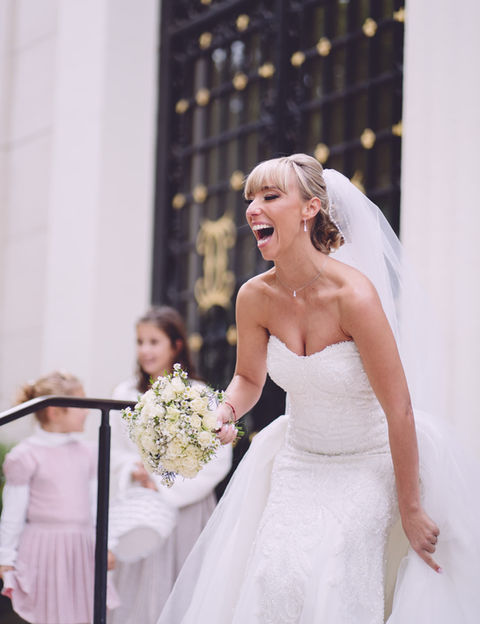 Mariée en robe blanche riant aux éclats, tenant un bouquet de fleurs blanches, entourée d’enfants à la sortie de la cérémonie de mariage.